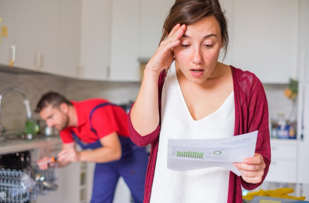 A woman in shock as she reads over the cost of her repair bill while a plumber works on her home appliance in the background