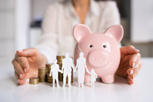 A woman sitting at a table with a piggy bank, a stack of coins, and a paper cutout of a family.