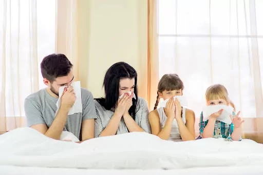 A dad, mom, and two children sititng on a bed and blowing their noses.