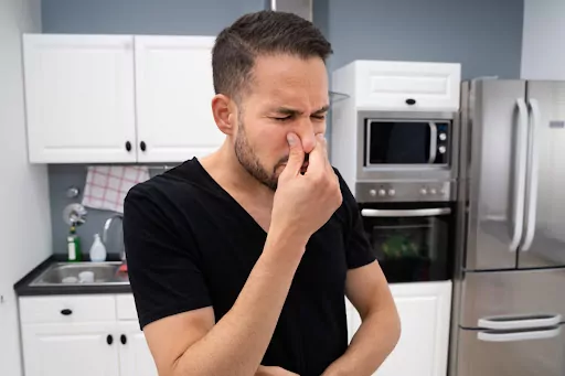 A man standing in a kitchen and holding his nose.