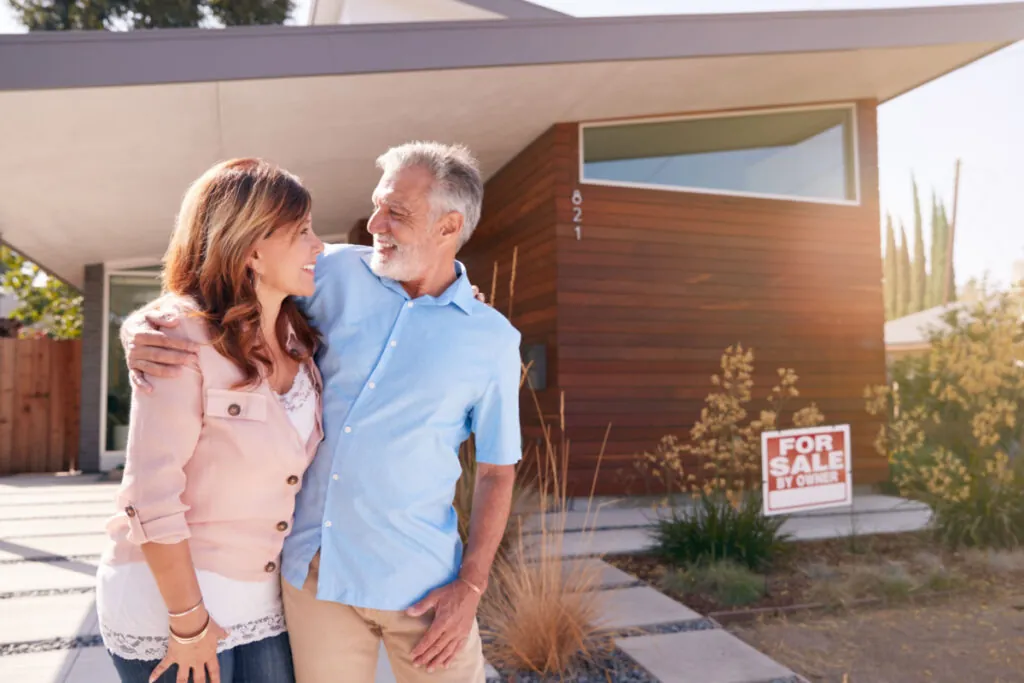 A smiling man and woman standing in front of a house with a "for sale" sign.